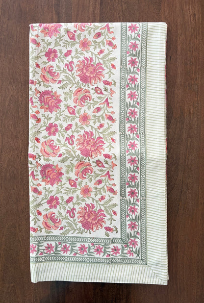 House of Mulmul - Blockprinted Cotton Table Cloth. A rectangular cream-colored textile, likely a tablecloth or napkin, featuring a vibrant pink and green floral pattern with a striped border, laid flat on a dark wood surface.