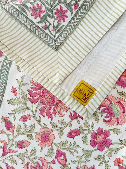 House of Mulmul - Blockprinted Cotton Table Cloth. A close-up of folded patterned fabric with three visible print styles: a green and cream striped border, a pink floral block print, and a pink floral vine pattern on a white base. A small gold fabric tag "House of Mulmul" is visible near the center.