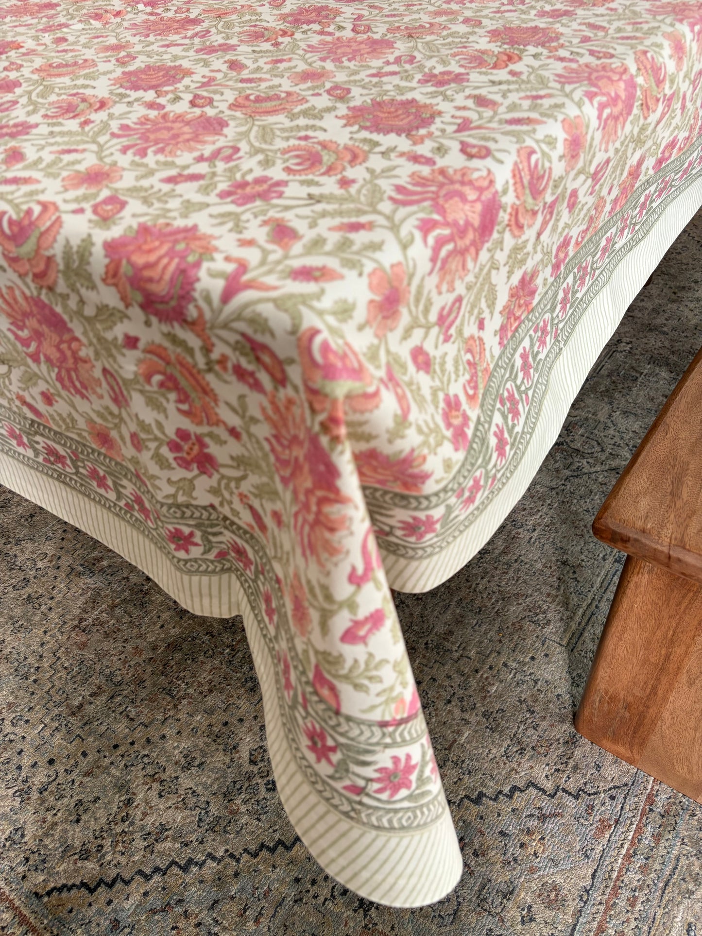 House of Mulmul - Blockprinted Cotton Table Cloth. A cream-colored tablecloth with a pink and green floral pattern and a striped border, draped over a table in a home setting next to a wooden stool.