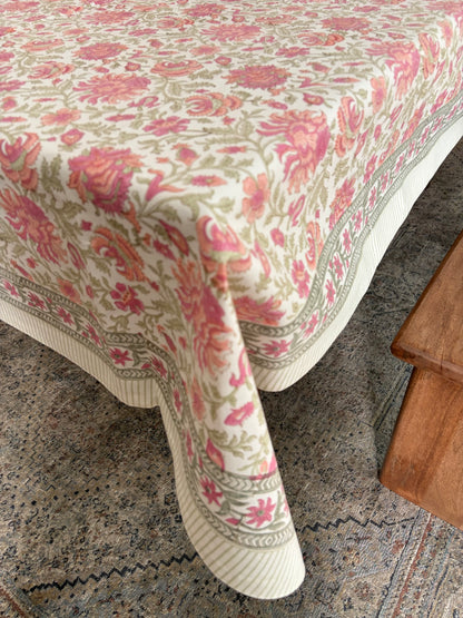 House of Mulmul - Blockprinted Cotton Table Cloth. A cream-colored tablecloth with a pink and green floral pattern and a striped border, draped over a table in a home setting next to a wooden stool.