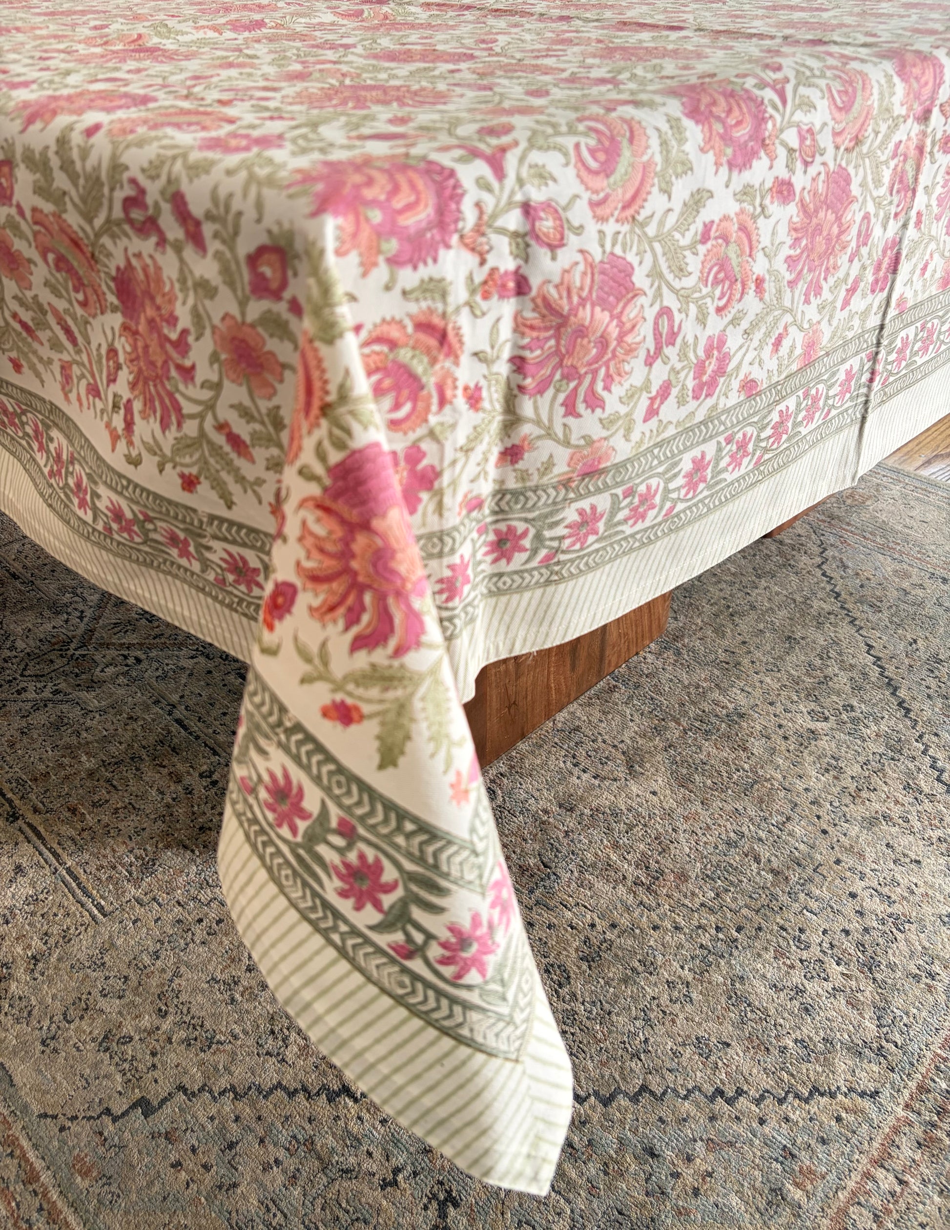 House of Mulmul - Blockprinted Cotton Table Cloth. A floral tablecloth draped over a table corner, featuring pink and red flowers with green leaves on an off-white background, set on a patterned rug.