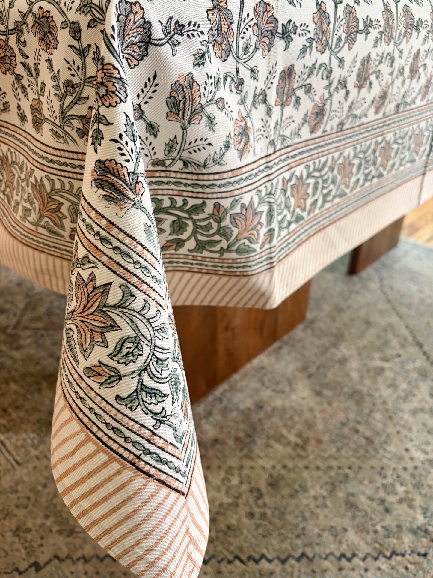 House of Mulmul - Blockprinted Cotton Table Cloth. A close-up of a patterned tablecloth draped over a wooden table, featuring a cream base with green and terracotta floral motifs and striped borders, set on a patterned rug.