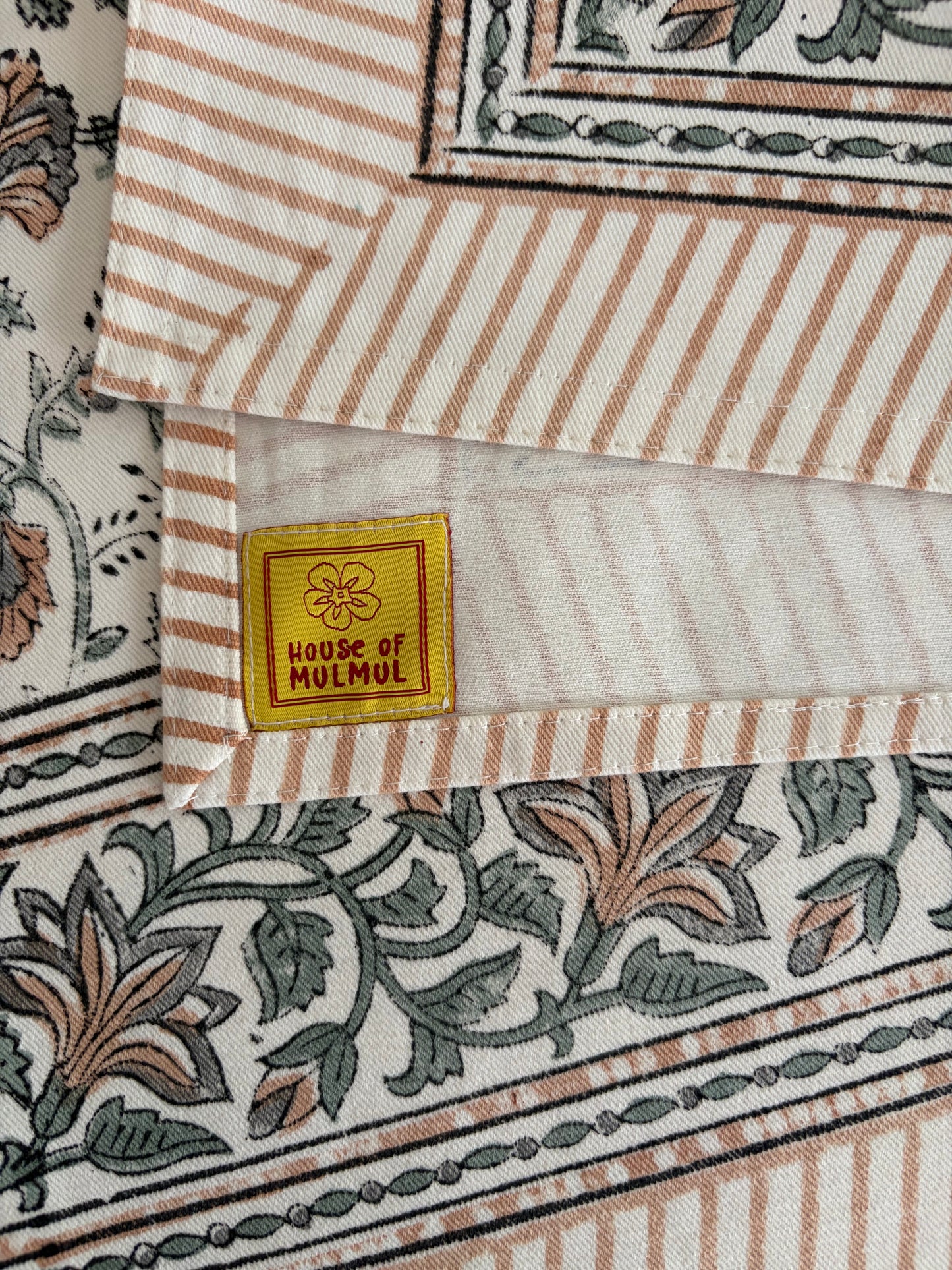 House of Mulmul - Blockprinted Cotton Table Cloth. A close-up of a folded textile product showing striped and floral printed fabric panels with a small yellow label reading 'HOUSE OF MULMUL'.