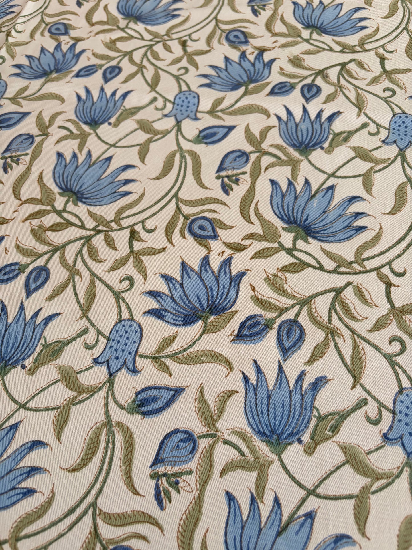 House of Mulmul - Blockprinted Cotton Table Cloth. A close-up of a fabric or wallpaper pattern featuring blue flowers and olive green vines on a warm cream background.