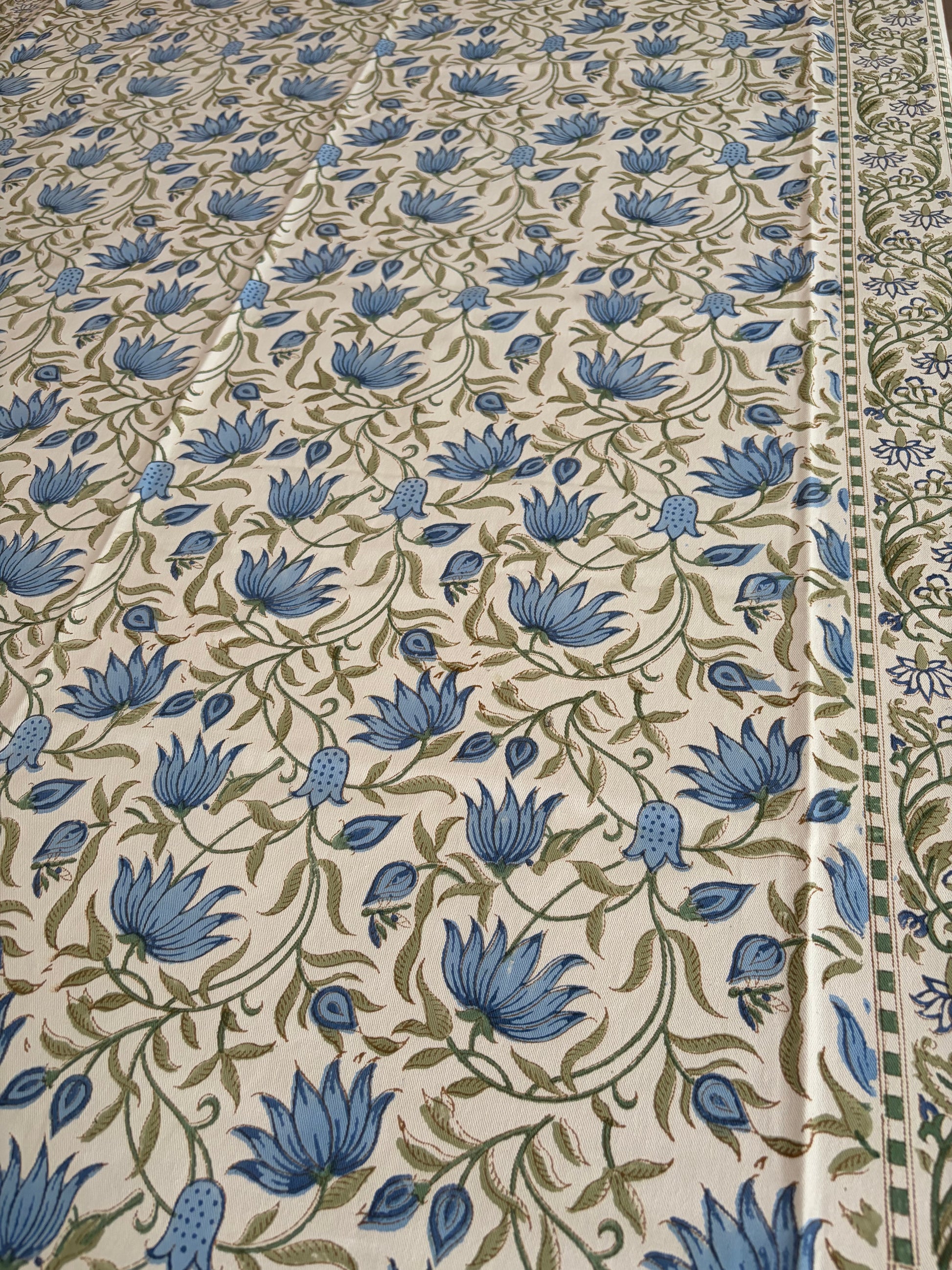 House of Mulmul - Blockprinted Cotton Table Cloth. A close-up of a printed textile or fabric with an allover floral pattern. The design features blue flowers and green vines on a warm cream background, with a decorative border visible along the right edge.