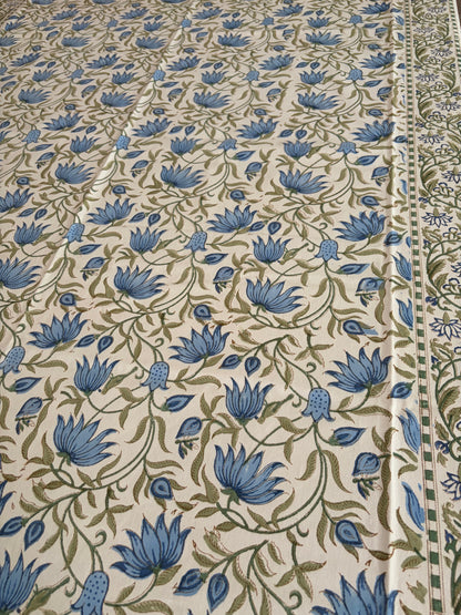 House of Mulmul - Blockprinted Cotton Table Cloth. A close-up of a printed textile or fabric with an allover floral pattern. The design features blue flowers and green vines on a warm cream background, with a decorative border visible along the right edge.