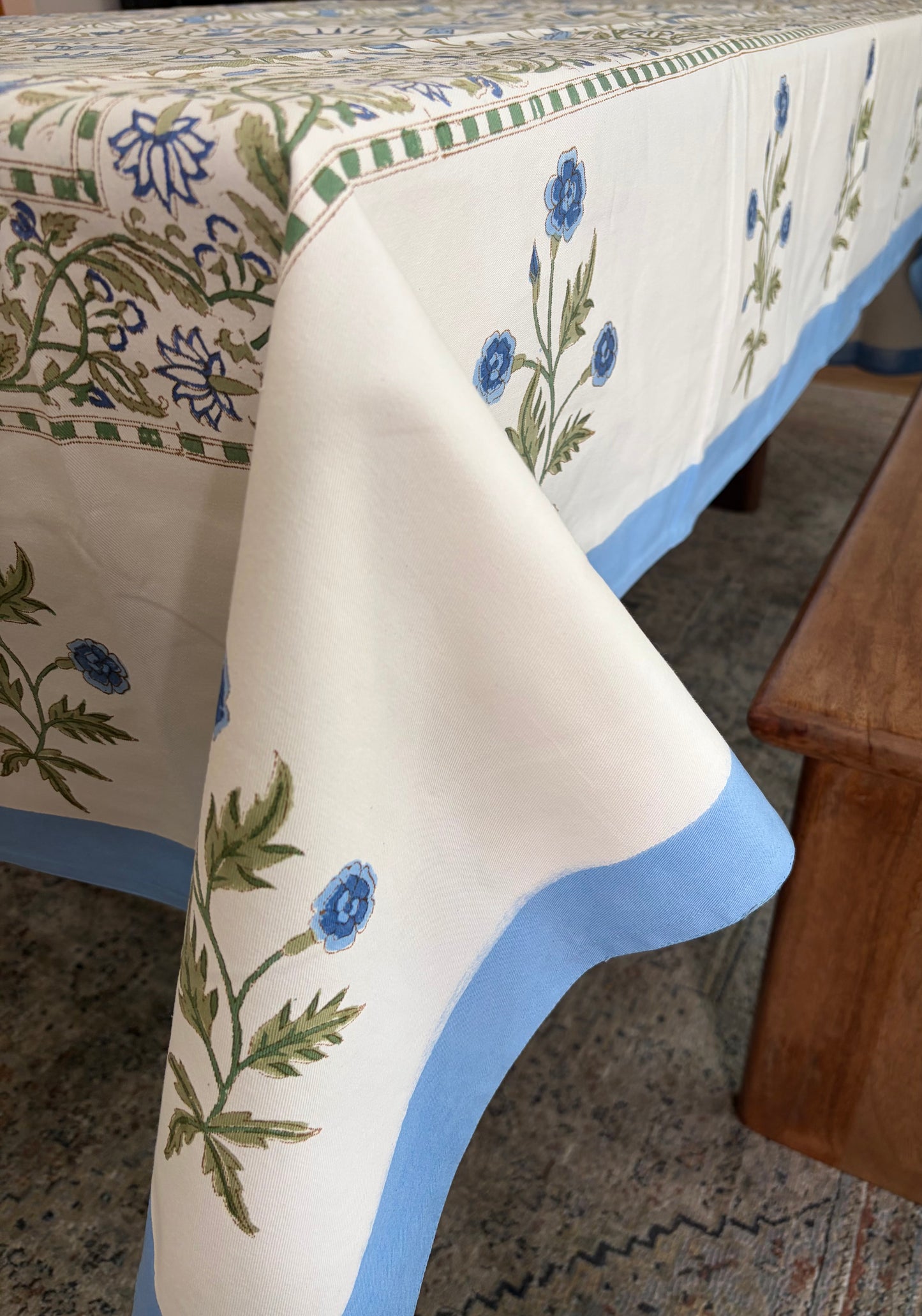 House of Mulmul - Blockprinted Cotton Table Cloth. A cream-colored cotton tablecloth with blue floral motifs and green leaves, finished with a light blue border, draped over a table in a home setting.