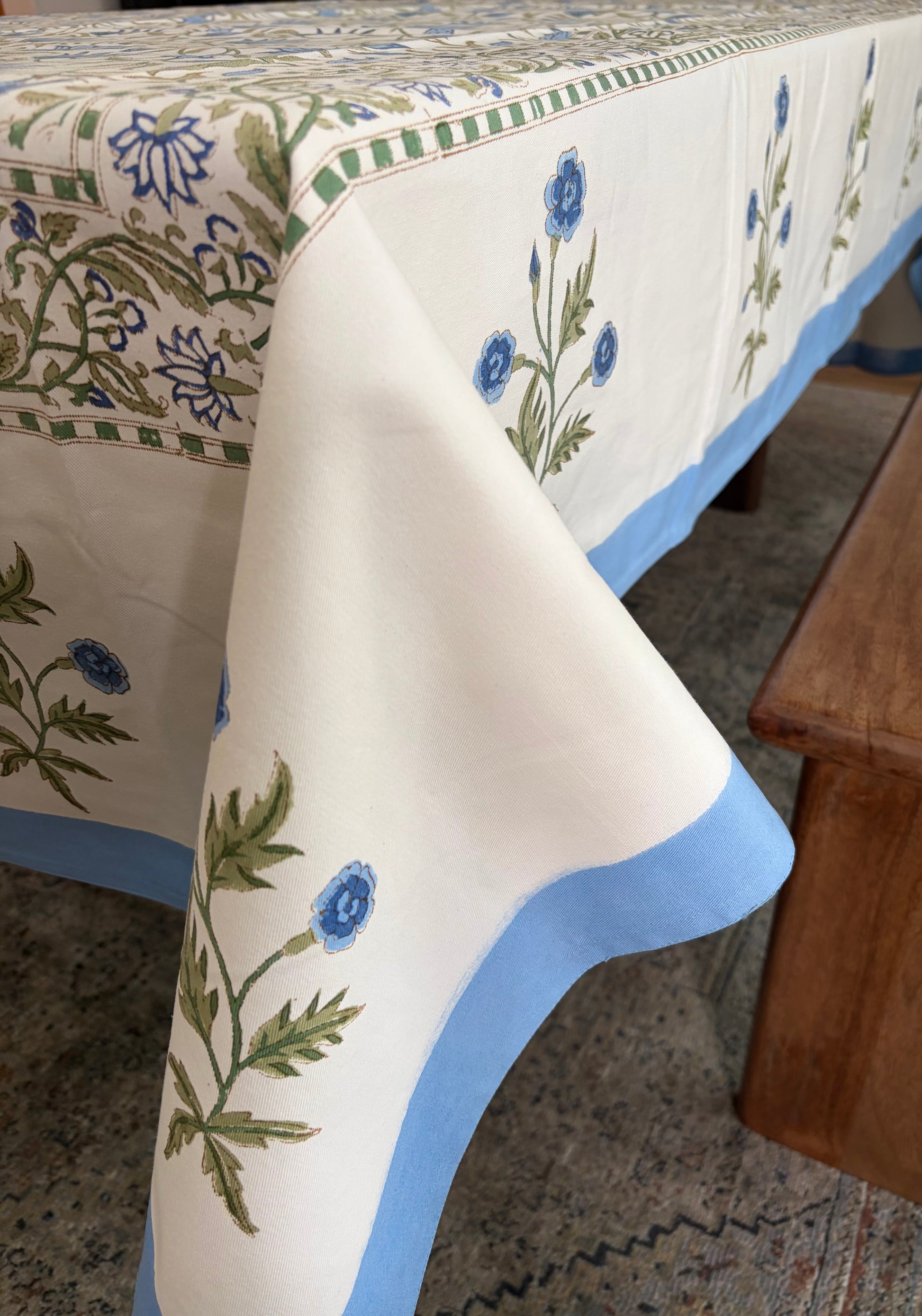 House of Mulmul - Blockprinted Cotton Table Cloth. A cream-colored cotton tablecloth with blue floral motifs and green leaves, finished with a light blue border, draped over a table in a home setting.