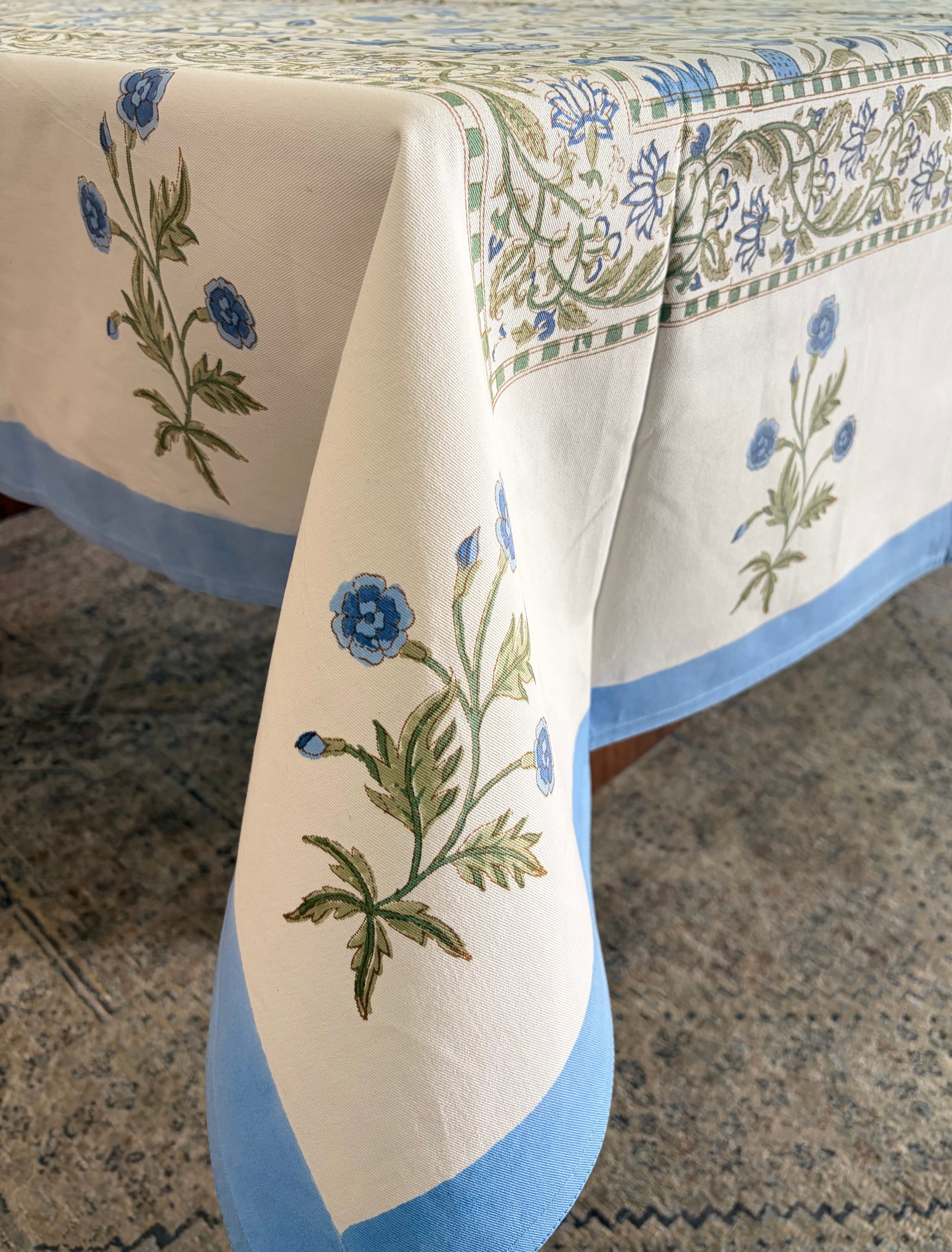 House of Mulmul - Blockprinted Cotton Table Cloth. A cream-colored cotton tablecloth with blue and green floral motifs and a solid blue border, draped over a table on a patterned rug.