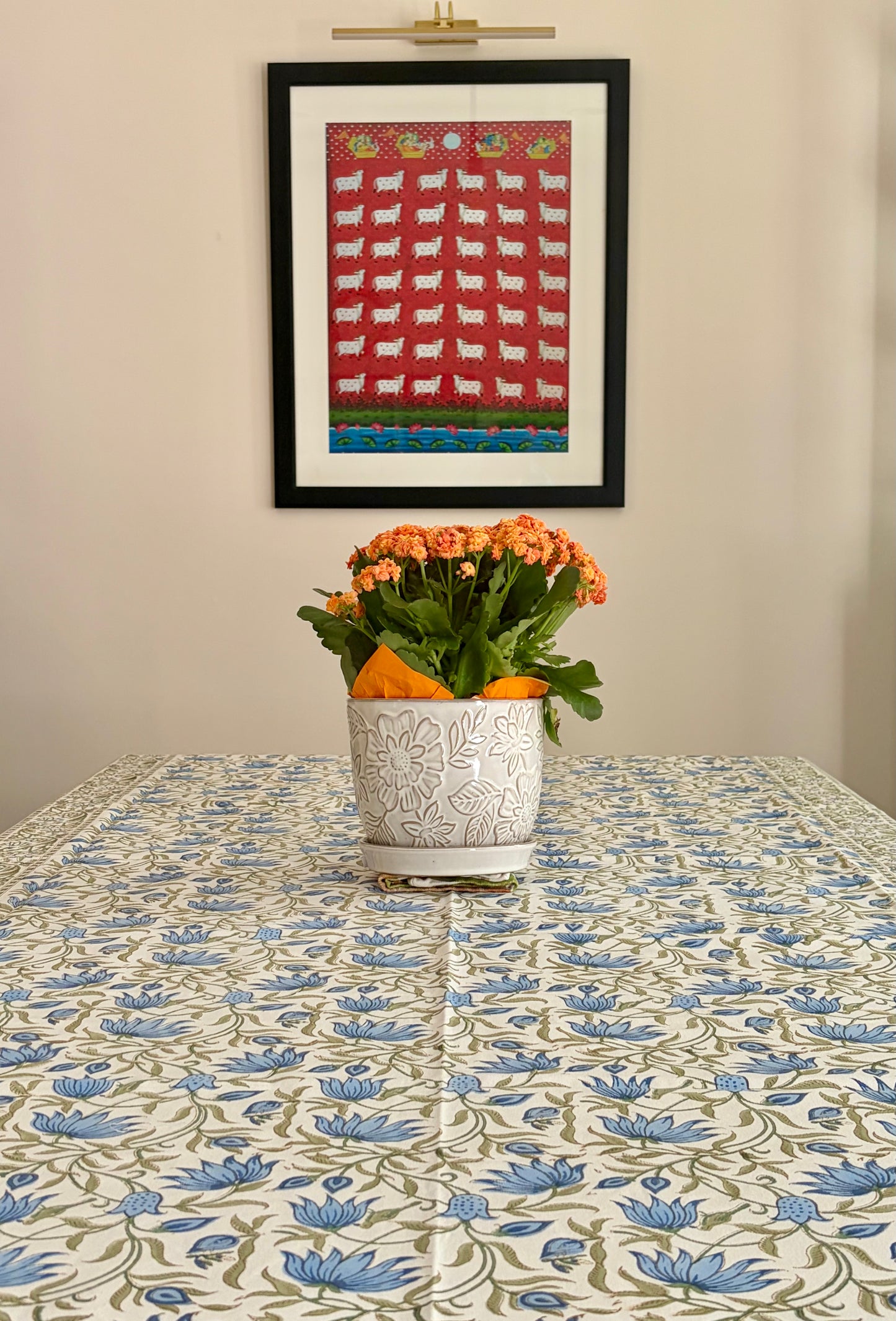 House of Mulmul - Blockprinted Cotton Table Cloth. A framed folk-art print featuring white sheep on a red background hangs on a beige wall above a table covered with a blue and green floral tablecloth. A white ceramic planter with orange flowers sits on the table.