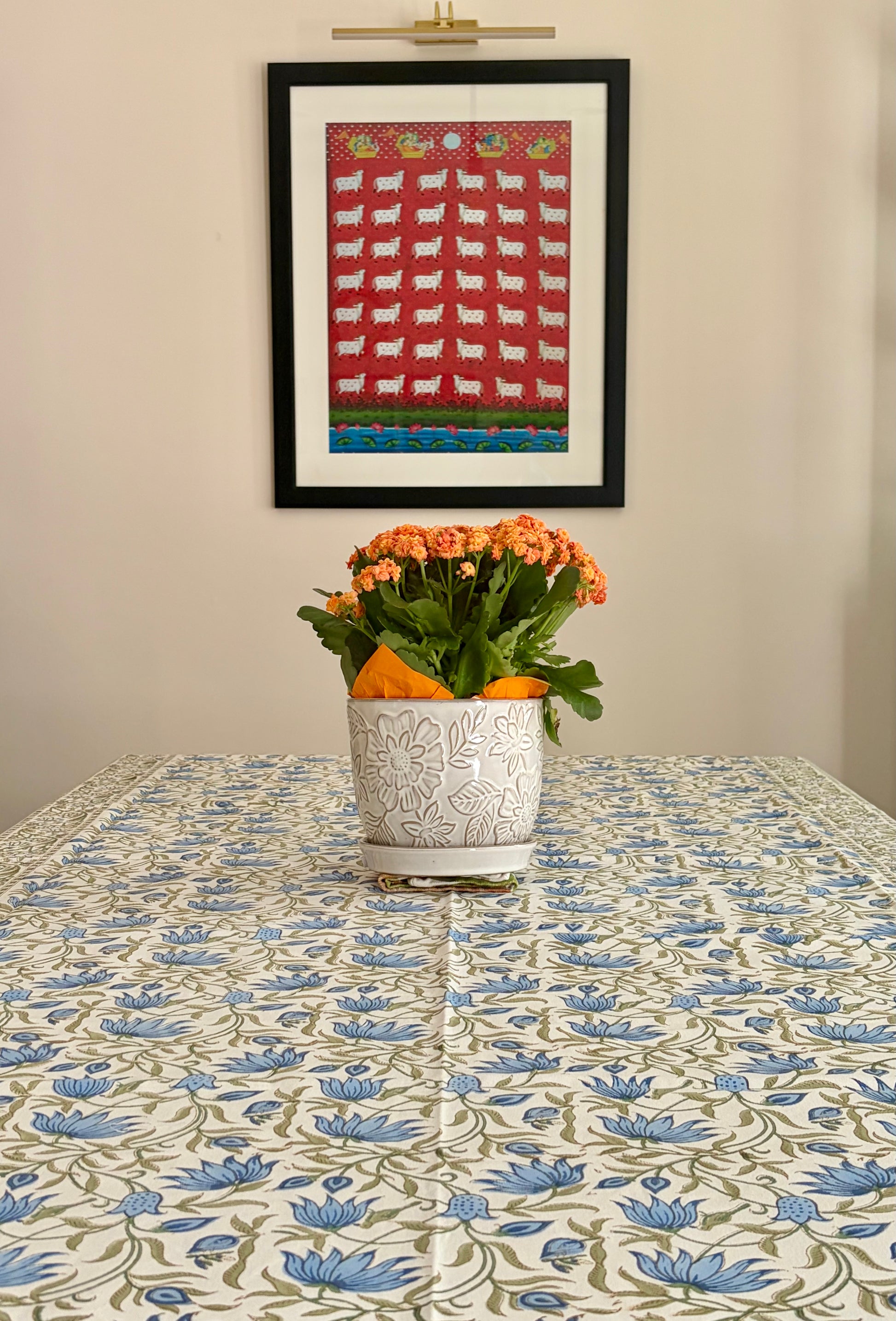 House of Mulmul - Blockprinted Cotton Table Cloth. A framed folk-art print featuring white sheep on a red background hangs on a beige wall above a table covered with a blue and green floral tablecloth. A white ceramic planter with orange flowers sits on the table.