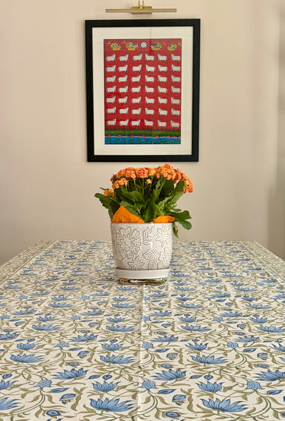 House of Mulmul - Blockprinted Cotton Table Cloth. A framed folk-art print featuring white sheep on a red background hangs on a beige wall above a table covered with a blue and green floral tablecloth. A white ceramic planter with orange flowers sits on the table.