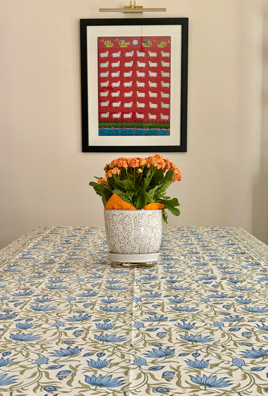 House of Mulmul - Blockprinted Cotton Table Cloth. A framed folk-art print featuring white sheep on a red background hangs on a beige wall above a table covered with a blue and green floral tablecloth. A white ceramic planter with orange flowers sits on the table.