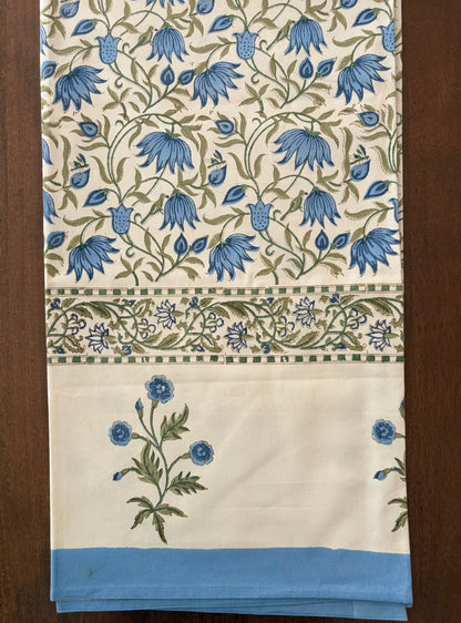 House of Mulmul - Blockprinted Cotton Table Cloth. A folded textile piece, likely a tablecloth or napkin, featuring a cream background with a blue and green floral vine pattern, a decorative border, and a solid light blue band at the bottom, displayed on a dark wood surface.