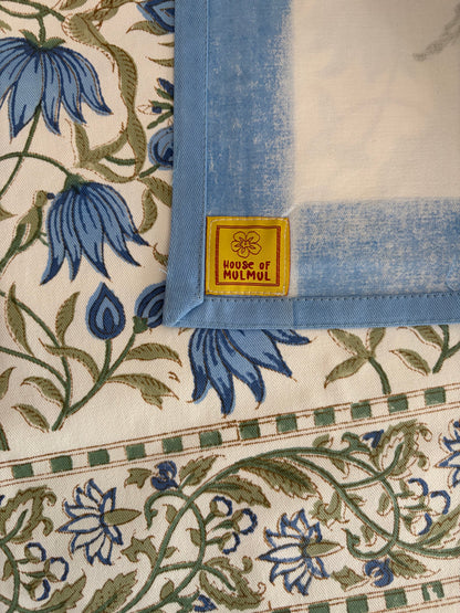 House of Mulmul - Blockprinted Cotton Table Cloth. A close-up of a House of Molmul textile, likely a table linen or napkin, showing a corner with a light blue border and a yellow brand label. The fabric features a cream background with a blue floral pattern and a green checkered border.
