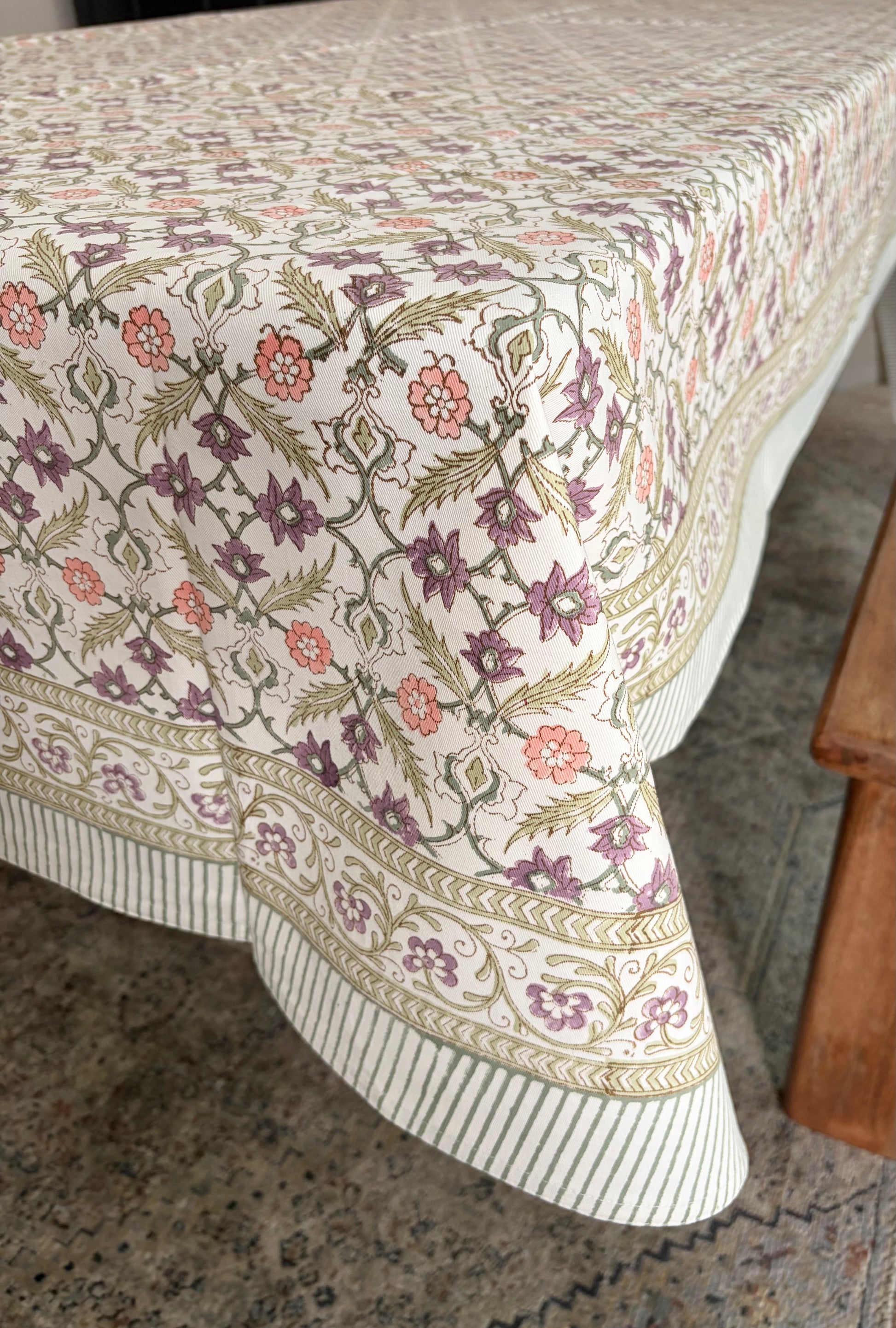 House of Mulmul - Blockprinted Cotton Table Cloth. A rectangular tablecloth with a detailed botanical and floral pattern in muted purple, orange, and olive green on an off-white background, draped over a table with a striped gathered border.