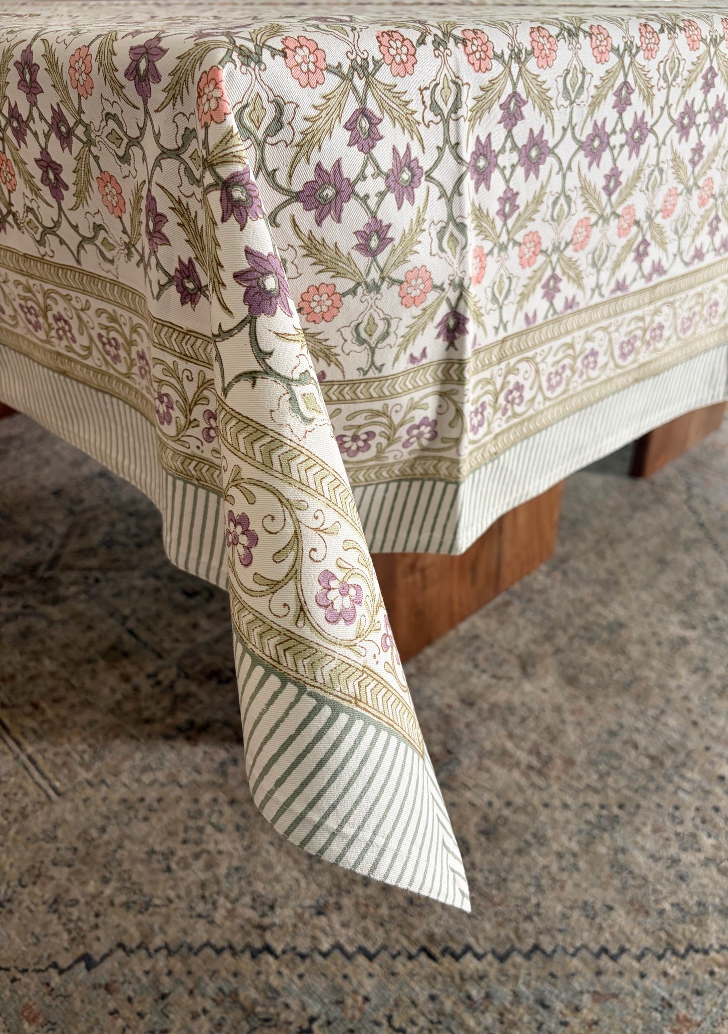 House of Mulmul - Blockprinted Cotton Table Cloth. A rectangular tablecloth with an intricate floral and botanical pattern in muted purple, green, and pink on an off-white background, draped over a table with a striped and patterned border.