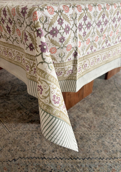 House of Mulmul - Blockprinted Cotton Table Cloth. A rectangular tablecloth with an intricate floral and botanical pattern in muted purple, green, and pink on an off-white background, draped over a table with a striped and patterned border.