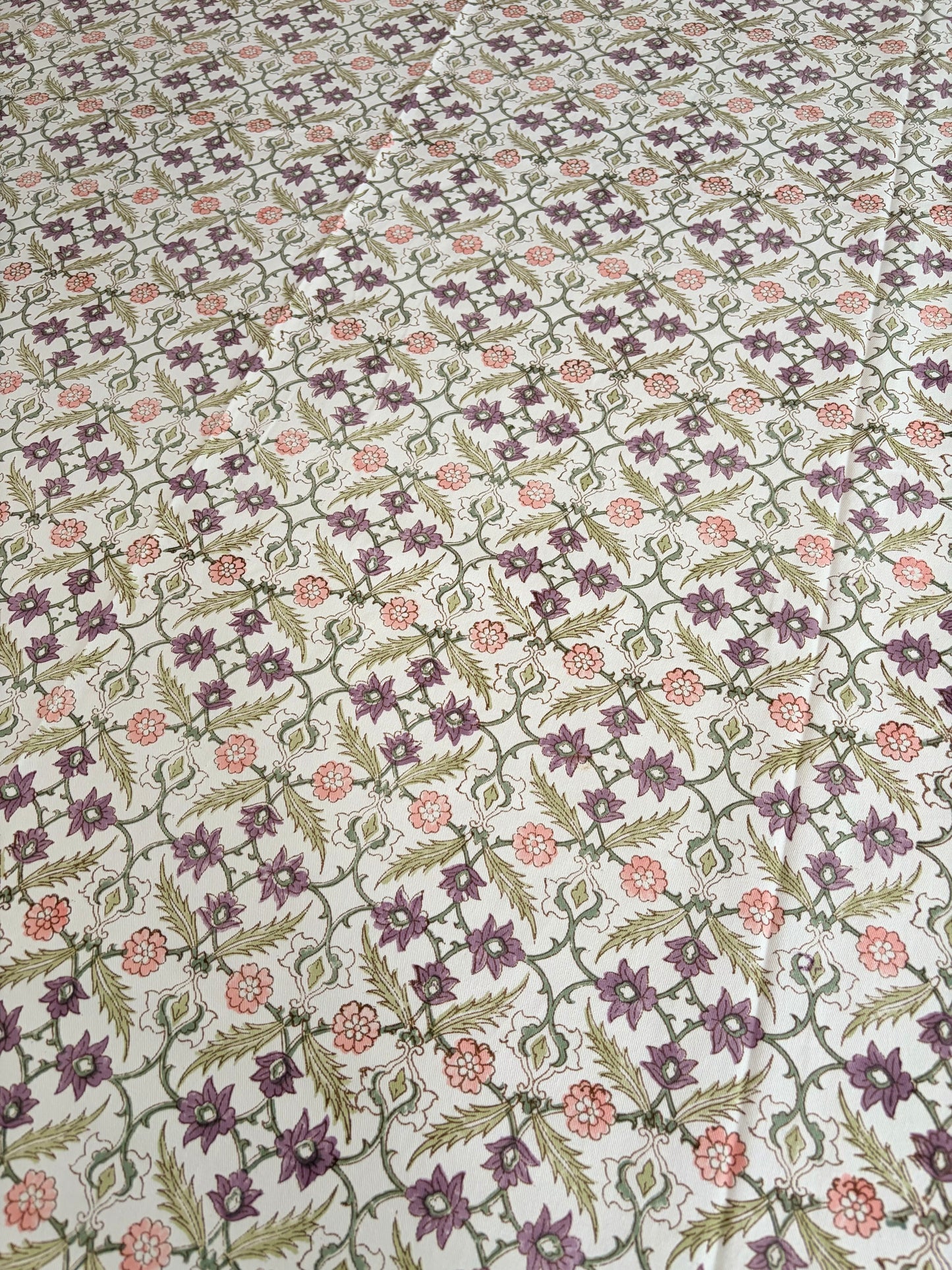 House of Mulmul - Blockprinted Cotton Table Cloth. A close-up of a fabric or wallpaper pattern featuring a repeating floral and geometric design in purple, green, and pink on a light cream background.