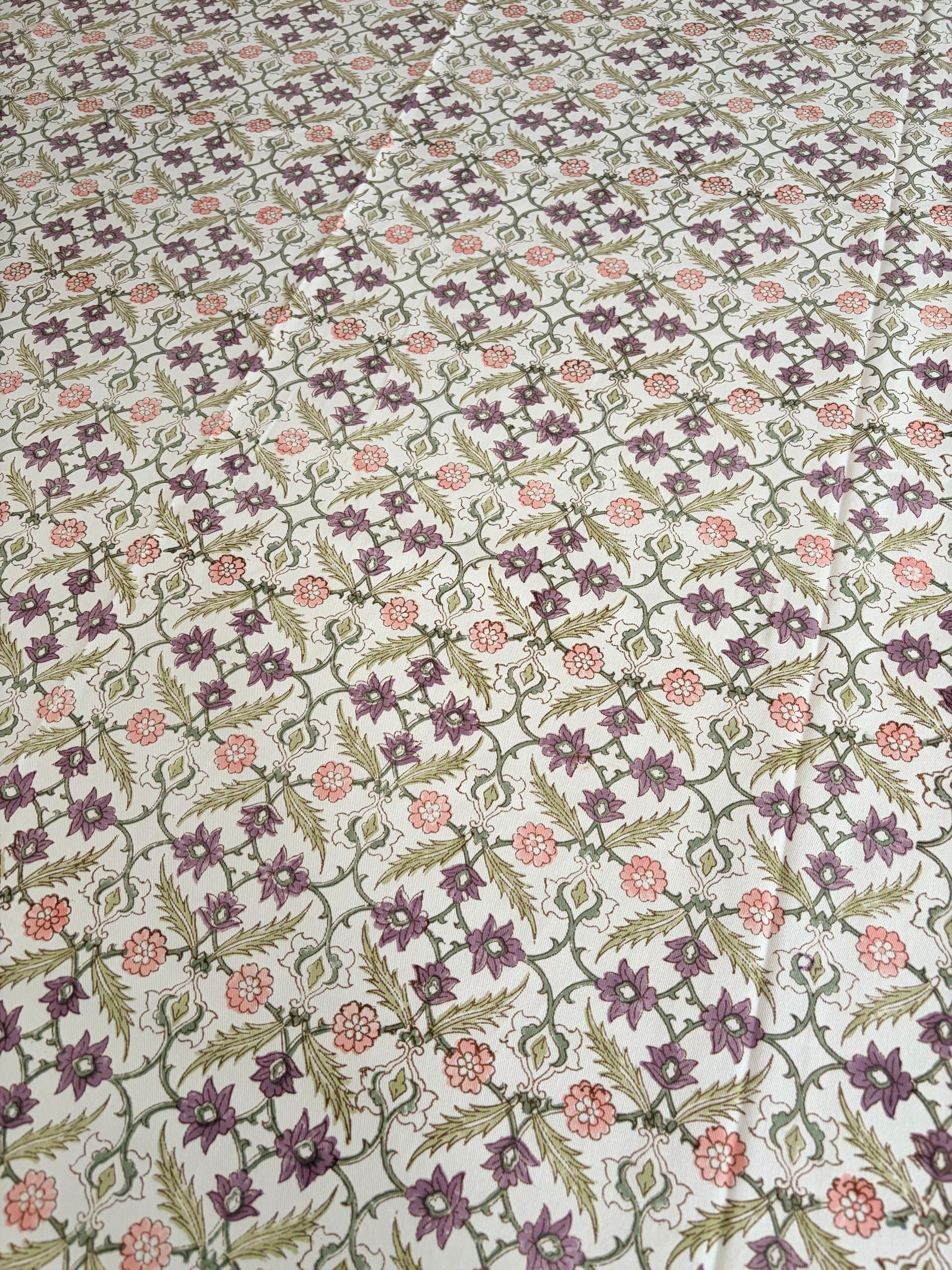 House of Mulmul - Blockprinted Cotton Table Cloth. A close-up of a fabric or wallpaper pattern featuring a repeating floral and geometric design in purple, green, and pink on a light cream background.