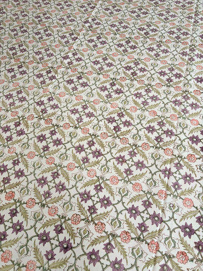 House of Mulmul - Blockprinted Cotton Table Cloth. A close-up of a fabric or wallpaper pattern featuring a repeating floral and geometric design in purple, green, and pink on a light cream background.