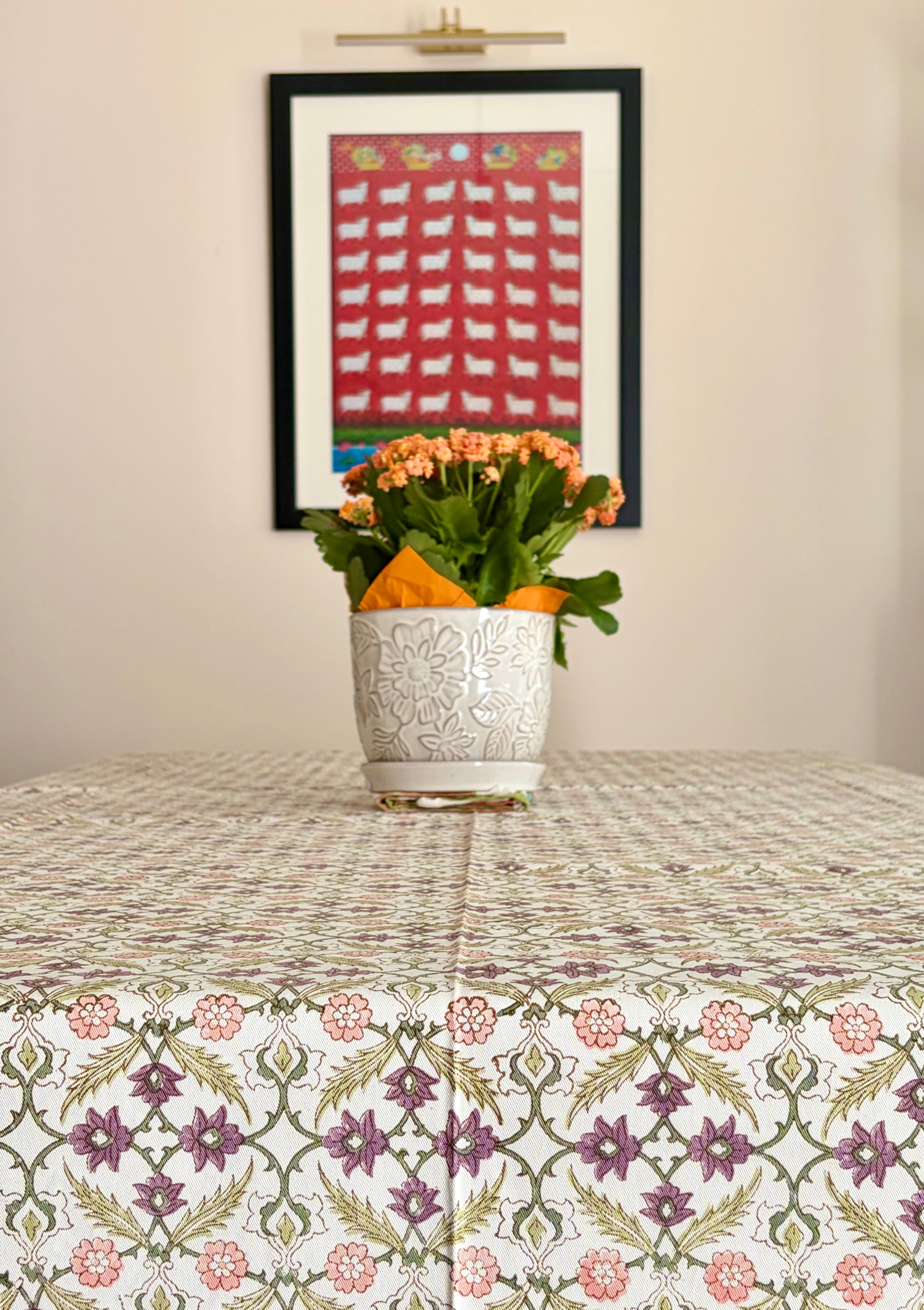House of Mulmul - Blockprinted Cotton Table Cloth. A patterned tablecloth with a floral and geometric design in muted pink, green, and purple tones, styled with a white textured ceramic vase of orange flowers on a table. In the background, a framed art print hangs on a beige wall.