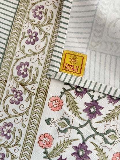 House of Mulmul - Blockprinted Cotton Table Cloth. Close-up of a folded textile product showing printed floral and geometric patterns in purple, pink, green, and beige, with a visible yellow label reading 'HOUSE OF MULMUL'.