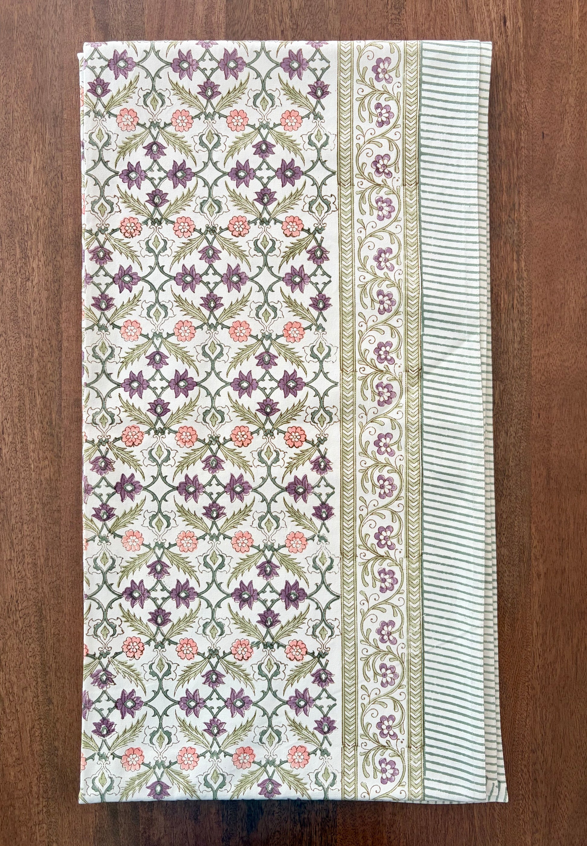 House of Mulmul - Blockprinted Cotton Table Cloth. A rectangular cotton table runner with an intricate block-printed floral pattern, laid flat on a dark brown wooden surface. The runner features a central panel with purple and pink flowers on a green and white geometric lattice, flanked by two borders with striped and floral details.
