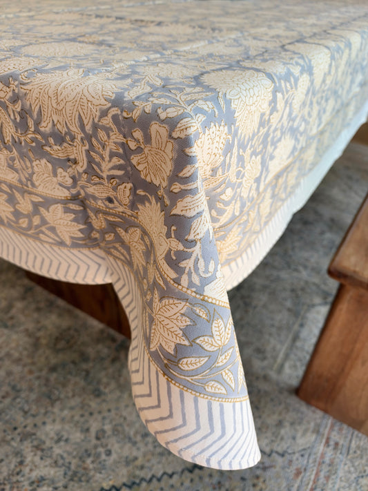 House of Mulmul - Blockprinted Cotton Table Cloth. A close-up of a tablecloth draped over a table, featuring a detailed beige floral and botanical pattern on a muted blue-gray background, with a striped border.