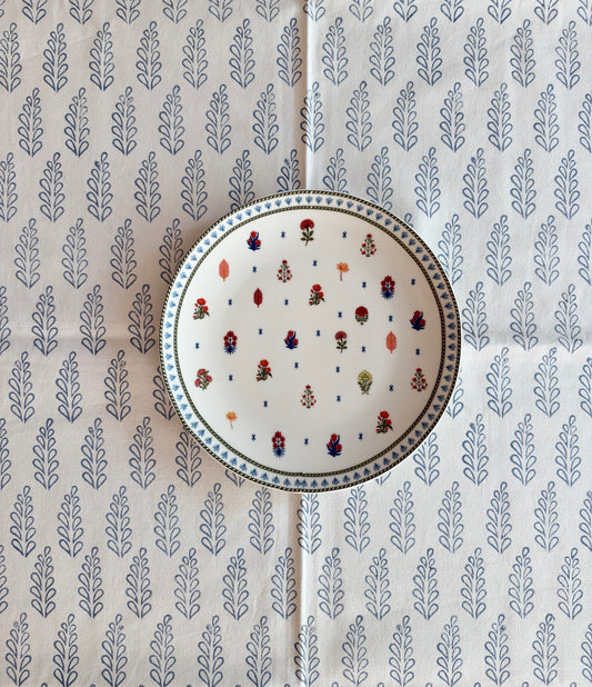 House of Mulmul - Blockprinted Cotton Tablecloth. A round white ceramic plate with a hand-painted block-print style floral pattern in red, blue, and green, placed on top of a matching patterned cream-colored tablecloth.
