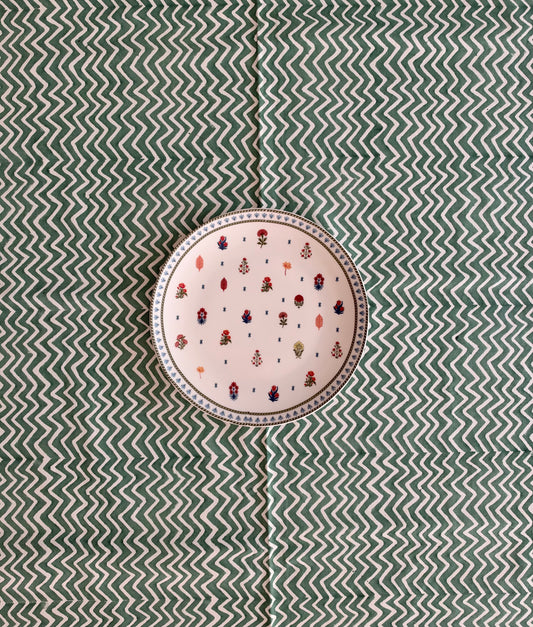 House of Mulmul - Blockprinted Cotton Tablecloth. A round ceramic plate with a light pink background and a pattern of small colorful birds, set against a green and white chevron-patterned fabric backdrop.