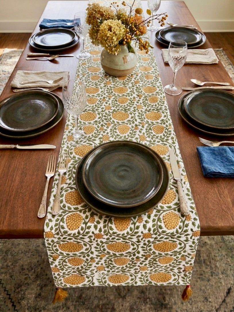 House of Mulmul - Blockprinted Cotton Reversible Table Runner & Placemats set. Dining table set with patterned table runner, dark plates, and floral centerpiece.