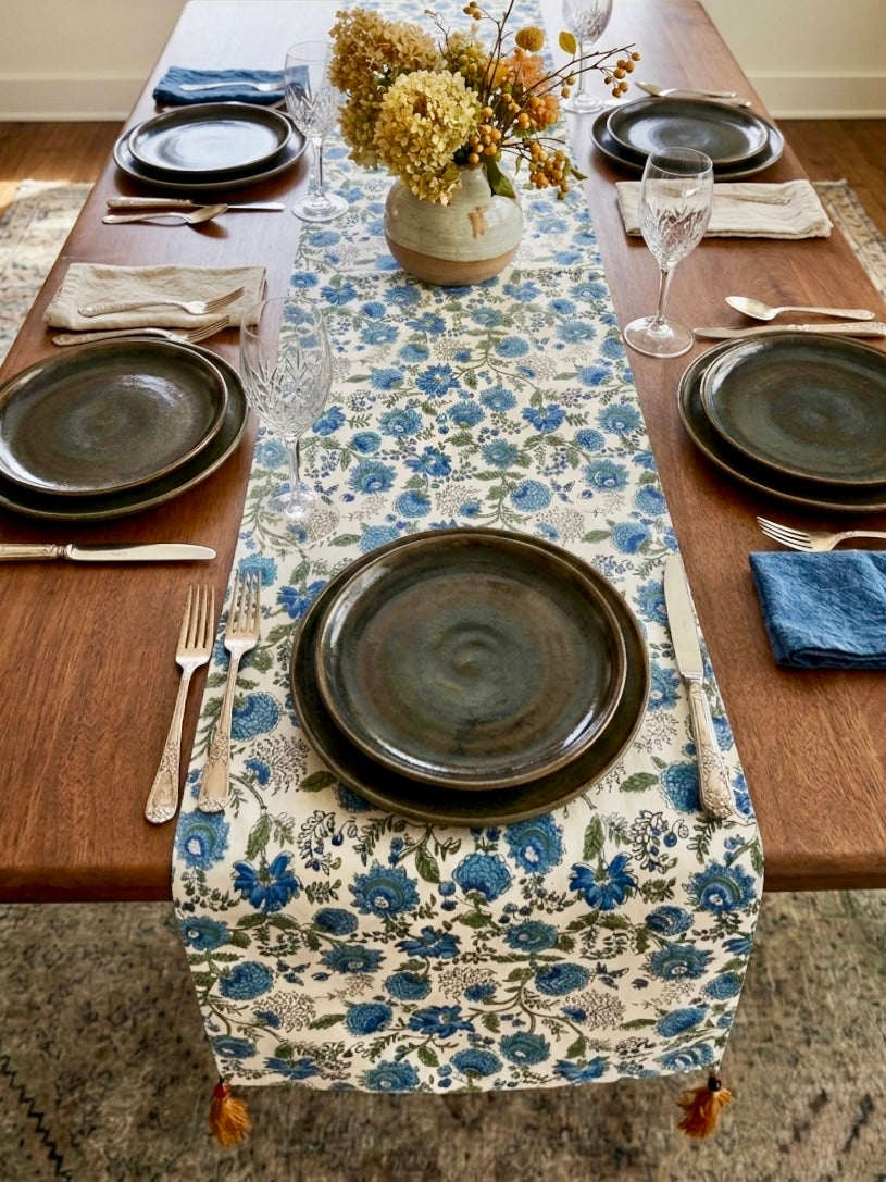 House of Mulmul - Blockprinted Cotton Reversible Table Runner & Placemats set. Dining table set with floral table runner, plates, and cutlery on a wooden floor.