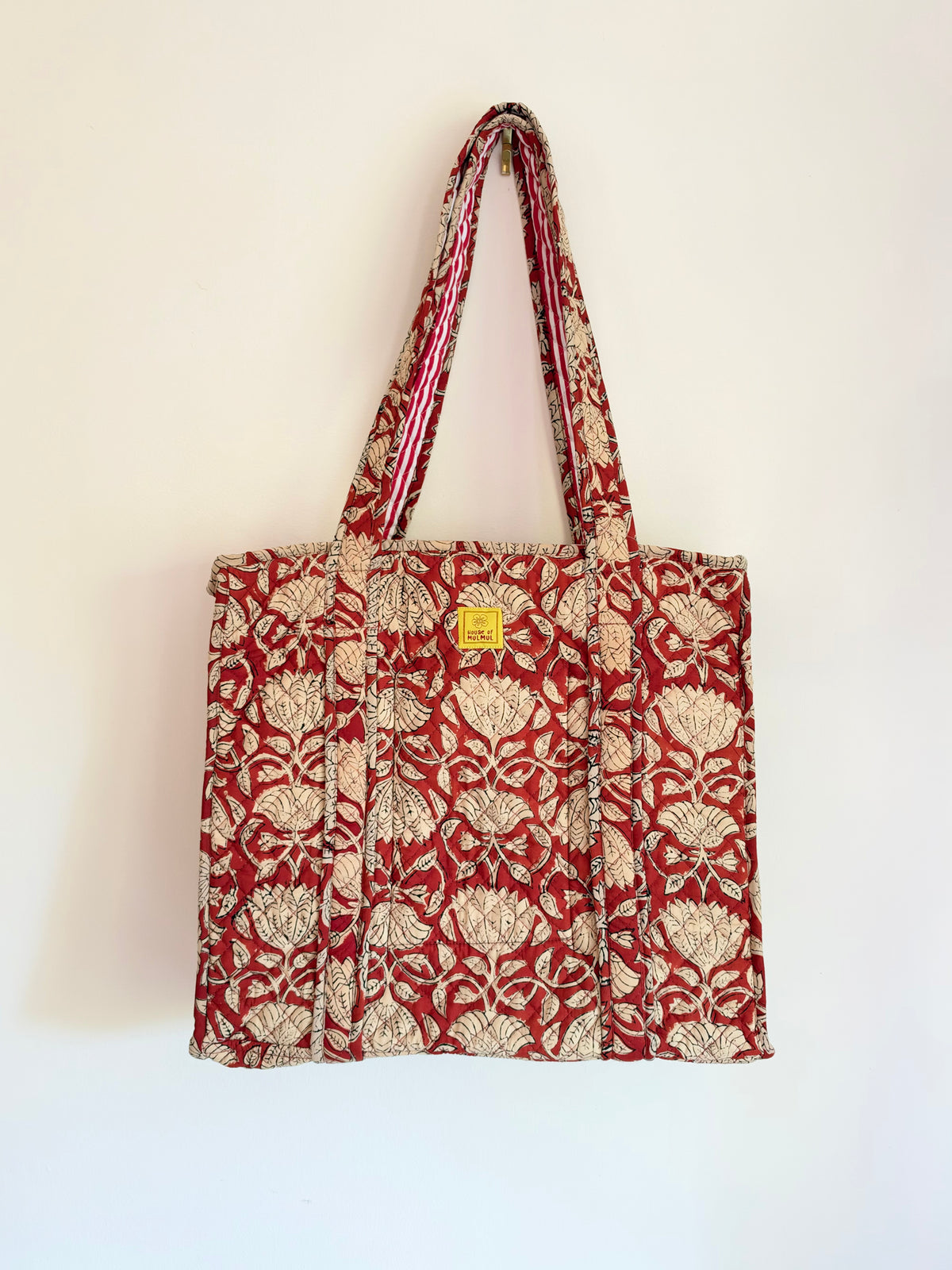 House of Mulmul - Blockprinted Cotton Tote Bags. A red and beige patterned canvas tote bag with matching shoulder straps, hanging against a plain off-white wall. The bag features a repeating floral motif and a small yellow square brand tag near the top center.