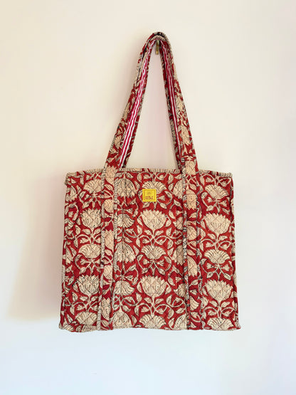 House of Mulmul - Blockprinted Cotton Tote Bags. A red and beige patterned canvas tote bag with matching shoulder straps, hanging against a plain off-white wall. The bag features a repeating floral motif and a small yellow square brand tag near the top center.