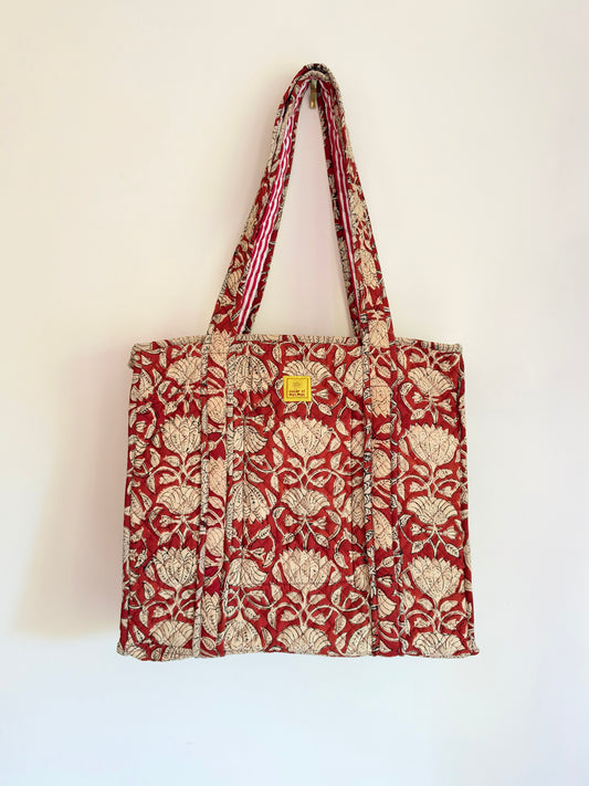 House of Mulmul - Blockprinted Cotton Tote Bags. A red and beige patterned canvas tote bag with matching shoulder straps, hanging against a plain off-white wall. The bag features a repeating floral motif and a small yellow square brand tag near the top center.