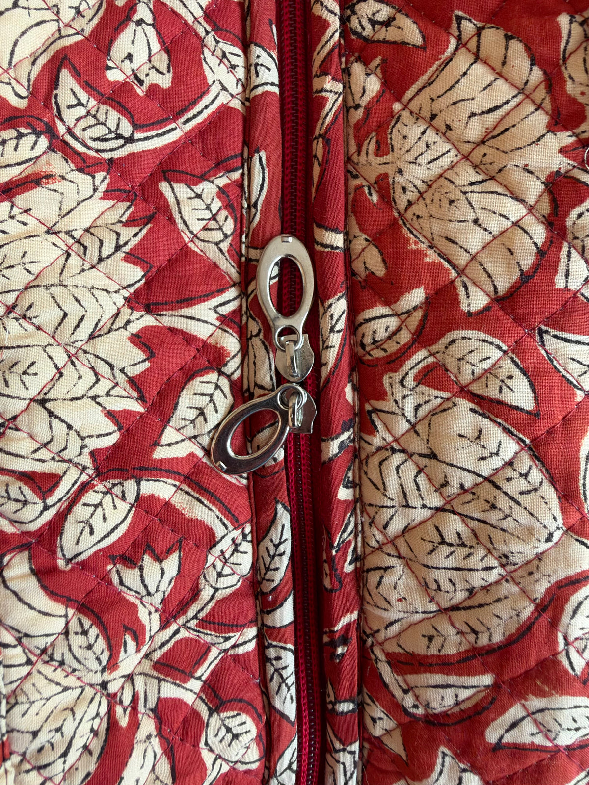 House of Mulmul - Blockprinted Cotton Tote Bags. Close-up of a quilted fabric jacket with a red and beige block-print leaf pattern, featuring a silver metal zipper and a matching red and beige leaf print.