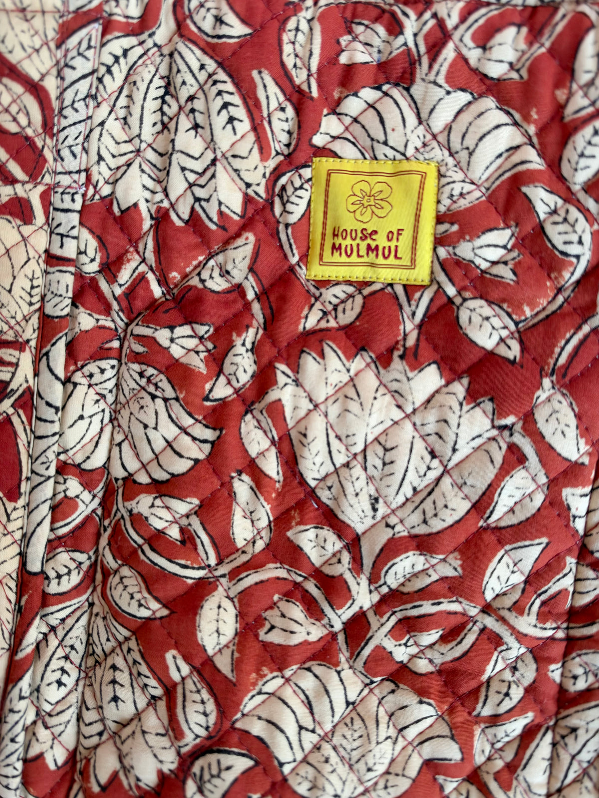 House of Mulmul - Blockprinted Cotton Tote Bags. A close-up of a quilted fabric featuring a red and off-white block-print floral and leaf pattern, with a yellow square brand label reading 'HOUSE OF MULMUL'.