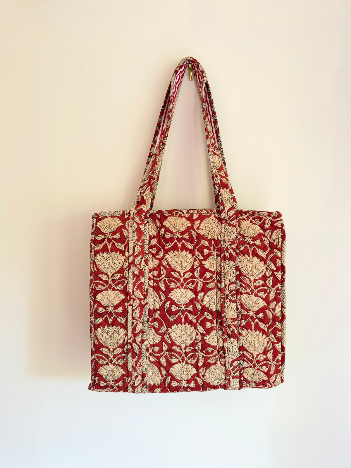 House of Mulmul - Blockprinted Cotton Tote Bags. A red and beige patterned fabric tote bag with long shoulder straps, hanging against a plain off-white wall.