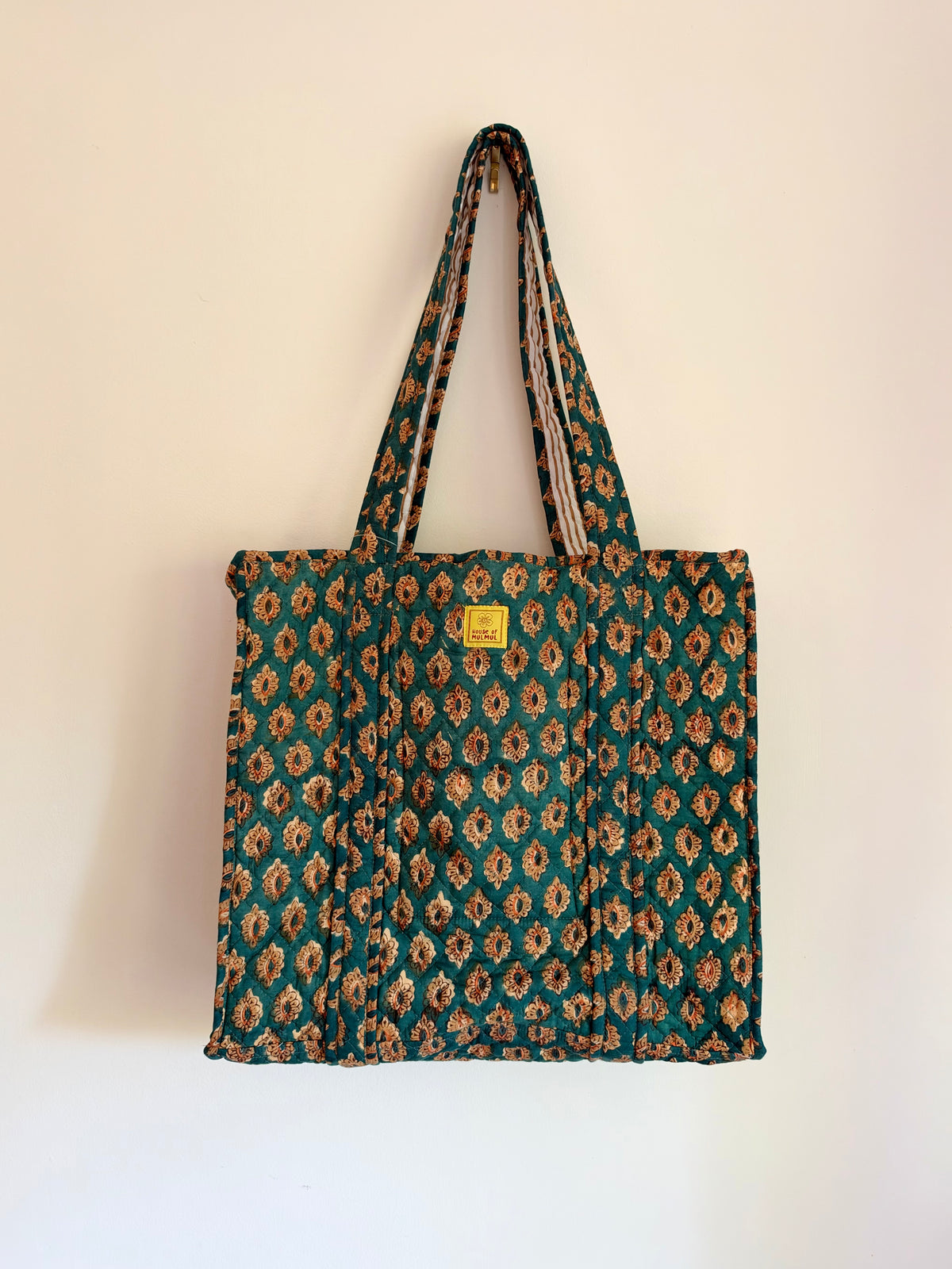 House of Mulmul - Blockprinted Cotton Tote Bags. A quilted fabric tote bag with a repeating floral ikat pattern in teal green, brown, and beige, featuring a small yellow brand label on the front, hanging against a plain off-white wall.