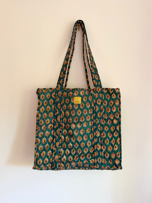 House of Mulmul - Blockprinted Cotton Tote Bags. A quilted fabric tote bag with a repeating floral ikat pattern in teal green, brown, and beige, featuring a small yellow brand label on the front, hanging against a plain off-white wall.