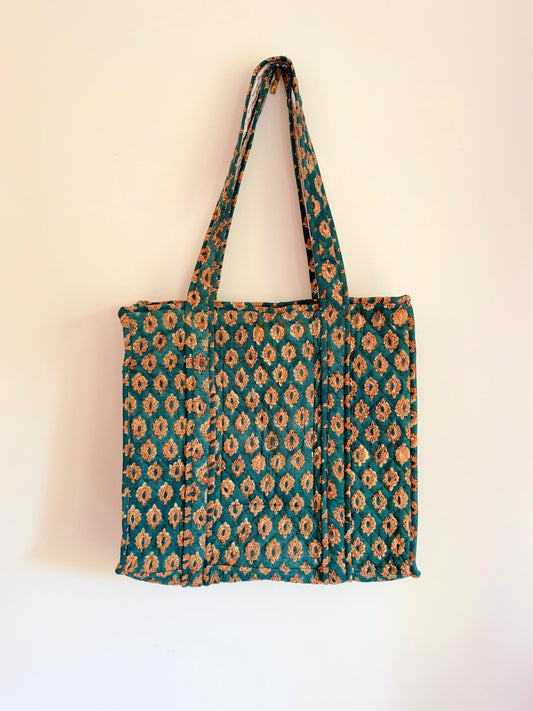 House of Mulmul - Blockprinted Cotton Tote Bags. A fabric tote bag with a teal green and beige geometric ikat pattern, hanging against a plain off-white wall.