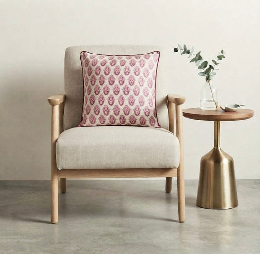 House of Mulmul - Blockprinted Cushion Covers-Pillow Cases. A light beige upholstered armchair with a pink patterned throw pillow, shown next to a round wooden side table with a gold metal base and a glass vase with greenery, against a neutral beige wall and gray concrete floor.