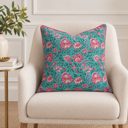 House of Mulmul - Blockprinted Cushion Covers-Pillow Cases. A square decorative throw pillow with a vibrant teal background and a pink and white floral pattern, styled on a cream-colored armchair next to a side table with a plant.
