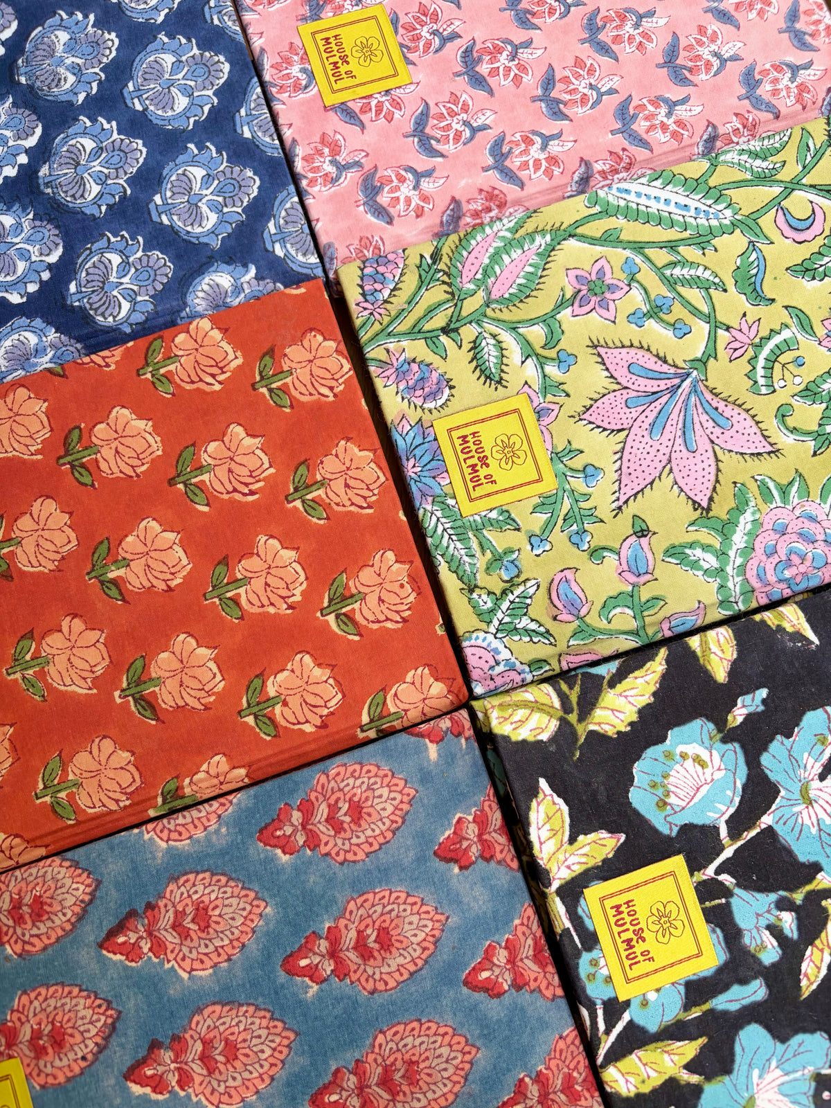 House of Mulmul - Blockprinted Journal Diary Notebook. Six patterned notebooks arranged in a grid, each with a different floral and botanical print in colors including blue, pink, orange, yellow, and black. Each sheet has a small yellow square label with text.