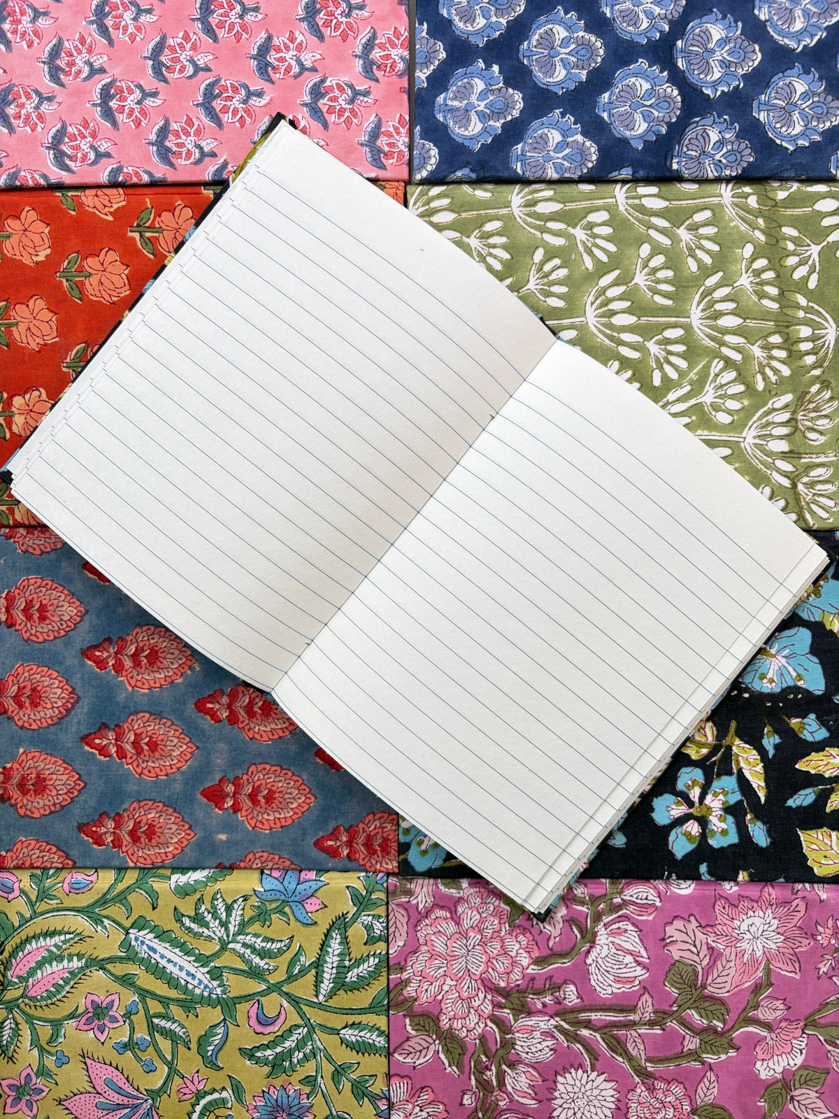 House of Mulmul - Blockprinted Journal Diary Notebook. An open lined notebook placed diagonally over a colorful patchwork background of patterned fabric swatches.