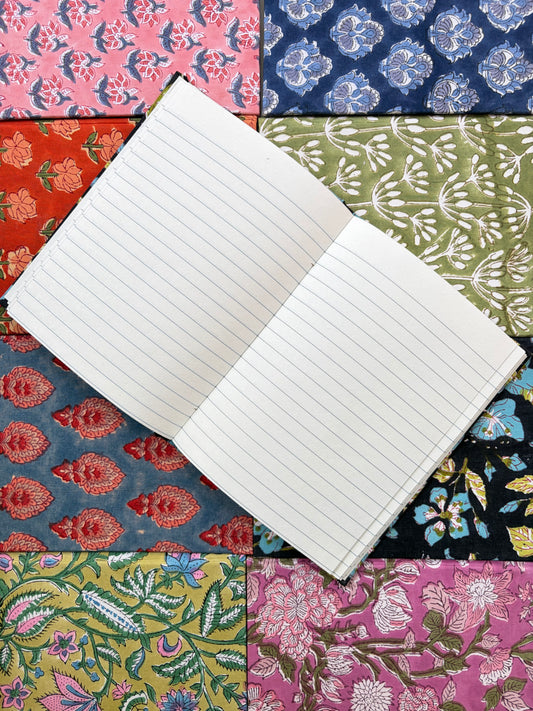 House of Mulmul - Blockprinted Journal Diary Notebook. An open lined notebook placed diagonally over a colorful patchwork background of patterned fabric swatches.