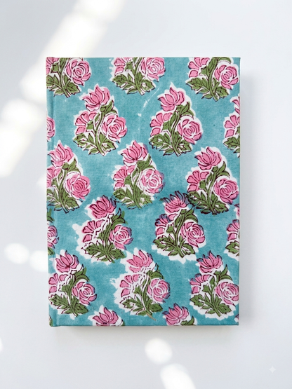 House of Mulmul - Blockprinted Journal Diary Notebook. A rectangular notebook featuring a repeating pink rose and green leaf block print on a turquoise blue background, displayed flat against a white surface with soft leaf shadows.