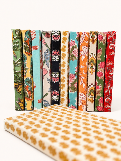 House of Mulmul - Blockprinted Journal Diary Notebook. A set of colorful patterned fabric-covered books standing upright, with a matching cream fabric-covered notebook or book in the foreground, all photographed on a clean white background.
