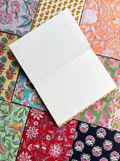 House of Mulmul - Blockprinted Journal Diary Notebook. An open blank notebook with white pages lies diagonally on top of a colorful patchwork arrangement of patterned paper sheets.